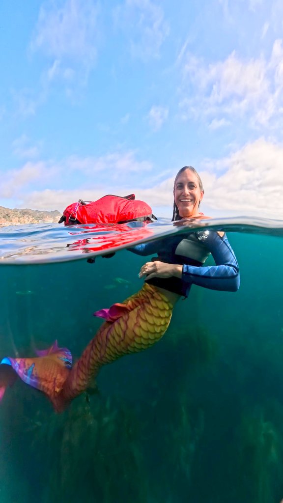 A PADI Mermaid Instructor swims in an orange mermaid tail while towing a red freediving buoy.